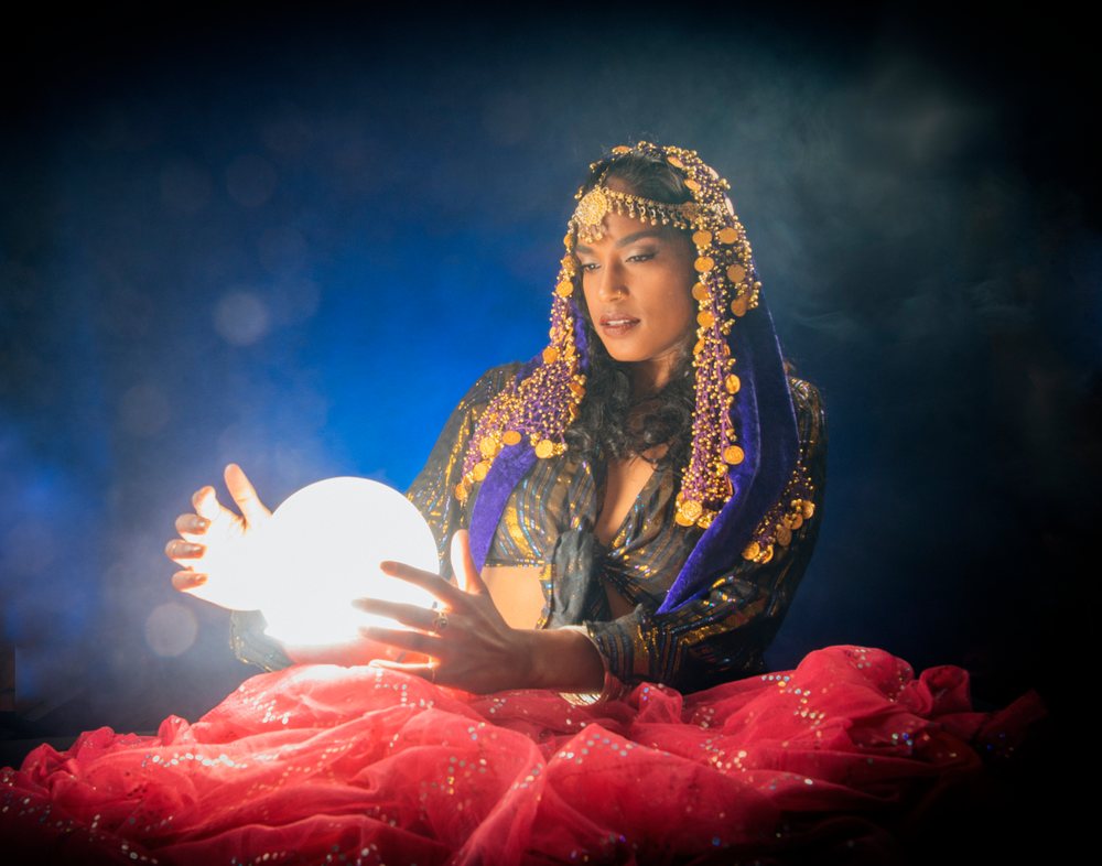 A woman wearing a purple headdress with gold coins holds a glowing orb indoors in the USA during a fortune telling scene, fortune teller, spirituality.