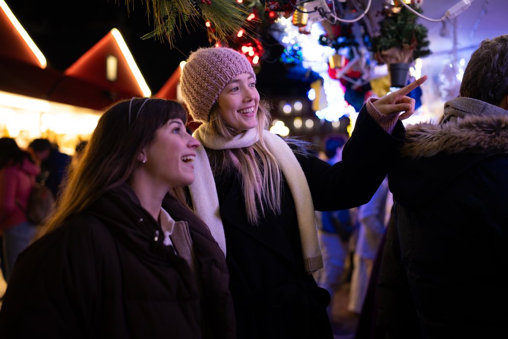 Two women smiling and pointing at festive lights at a Christmas market in Madrid at night, Spain.
