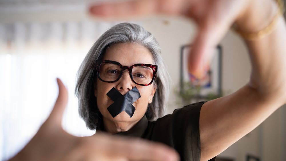 Middle-aged woman indoors with gray hair and glasses makes a framing gesture around her face, black tape covering her mouth symbolizing silence, Spain.