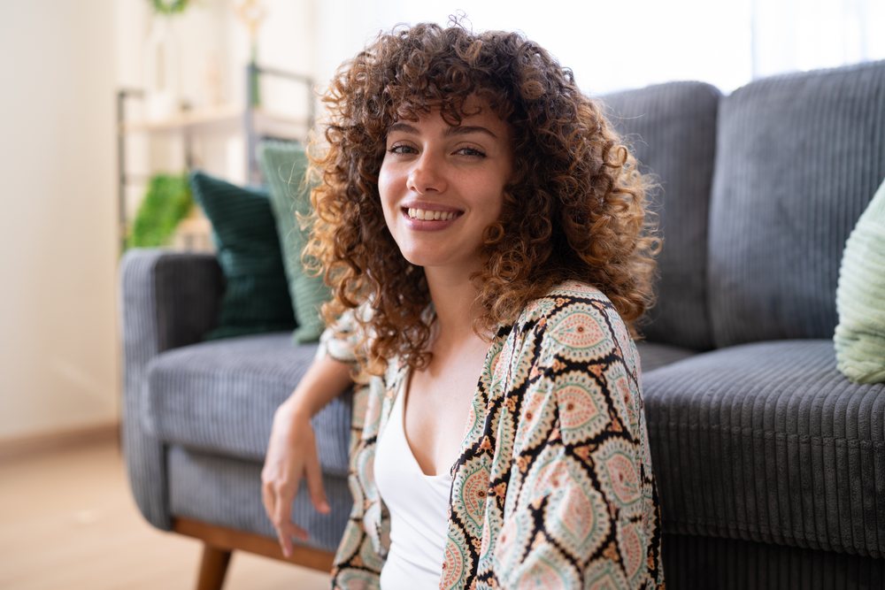 Woman with curly hair smiling while sitting indoors on the floor in a home living room with a gray couch and cushions behind her, Spain.