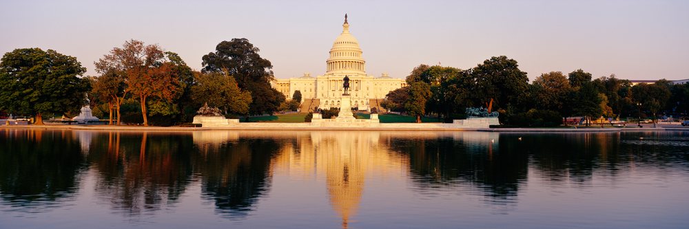 Sunset view of the United States Capitol building reflecting in the smooth water of the Capitol Reflecting Pool DC, with statues and trees nearby.