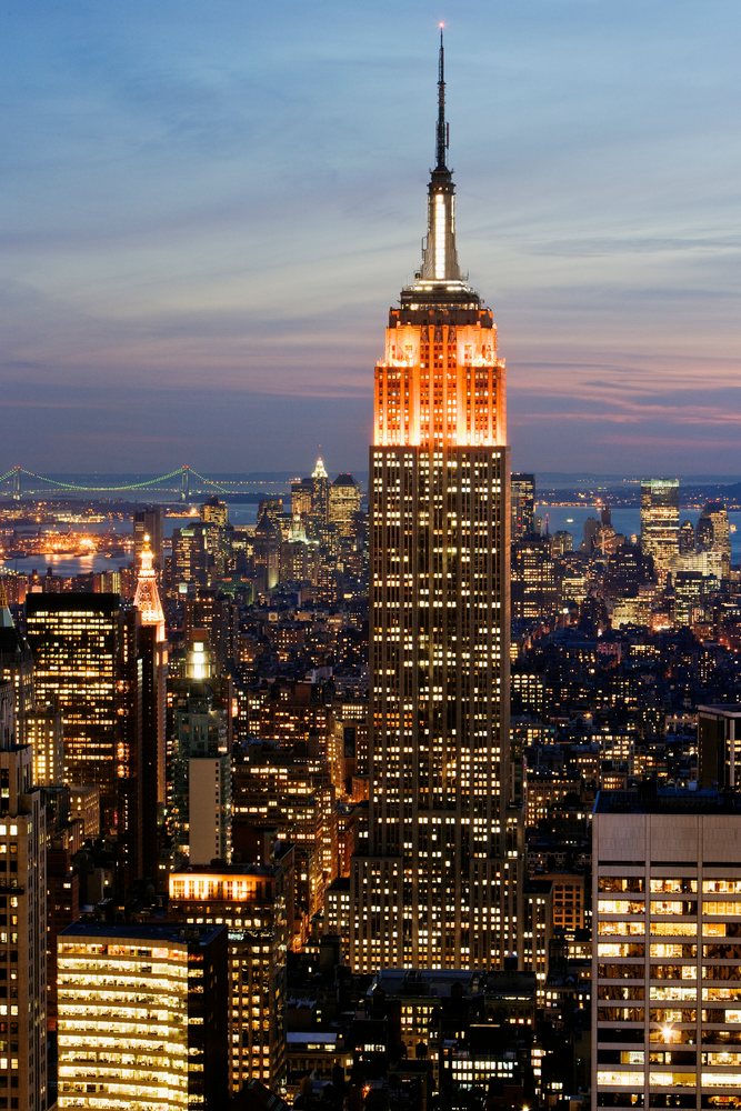 Empire State Building illuminated at twilight in the Manhattan skyline with surrounding skyscrapers and a river in view, New York, USA.