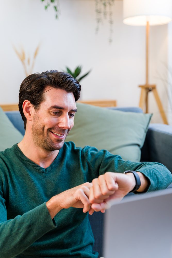Man smiling while checking his watch seated on a couch in a cozy indoor living room with plants and a floor lamp, Spain.