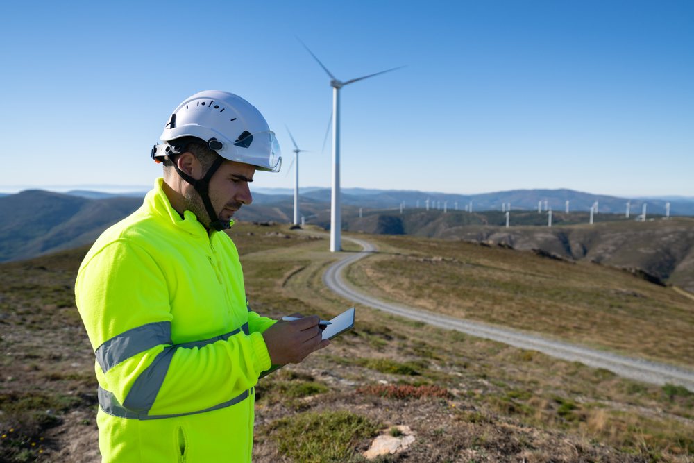 Man wearing safety helmet and neon high visibility jacket uses tablet at a wind farm, with wind turbines in the background, sustainability, renewable energy, Galicia, Spain.