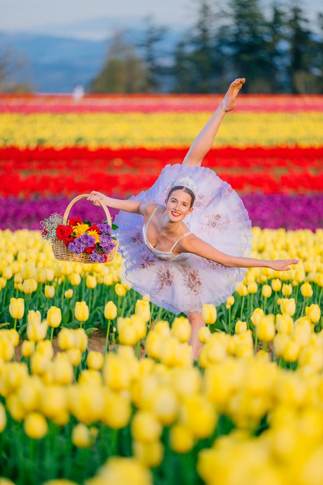 Ballet dancer wearing a tutu and headpiece balances on one leg holding a basket of assorted flowers in a colorful tulip field, USA.