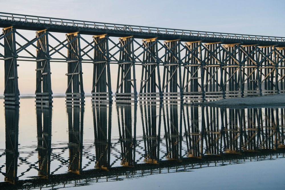 Wooden bridge with support beams reflecting in calm water at a beach during sunset, northern, Oregon, California, USA.
