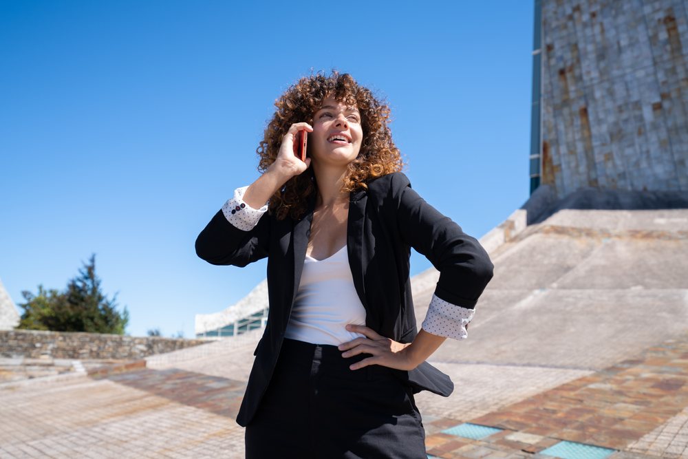 Businesswoman with curly hair talking on a mobile phone outdoors near modern architecture in Santiago de Compostela, traveling executive, Spain, Antiago De Compostela.