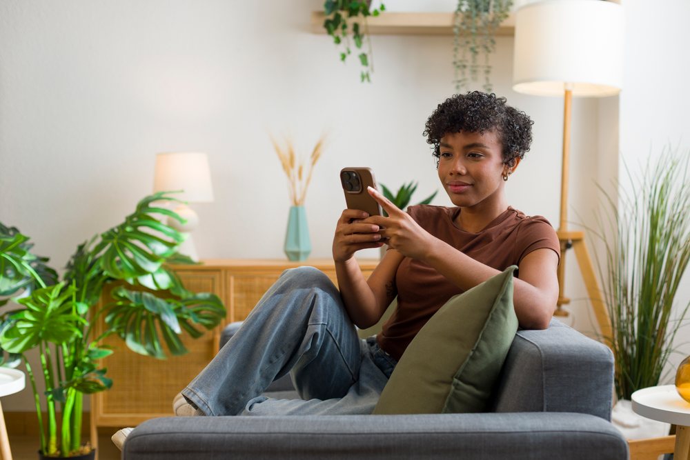 Young woman with curly hair sits on a gray sofa holding a smartphone in a modern living room with plants and wooden furniture.