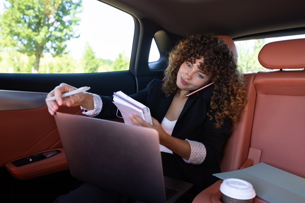 Woman multitasking by working remotely on a laptop and writing in a notebook inside a car, remote work, businesswoman, Spain.