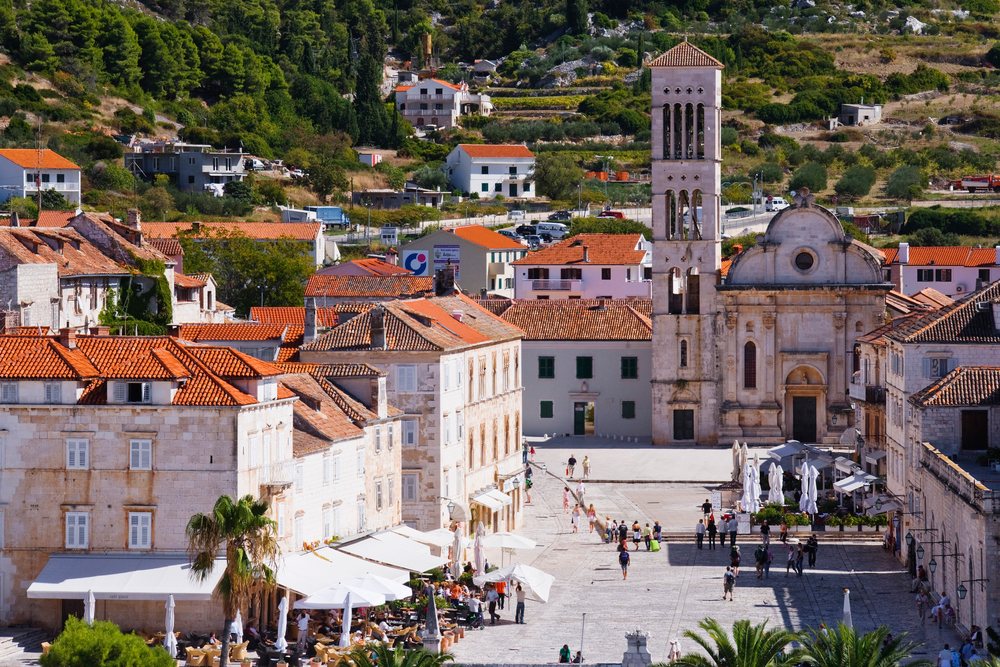 Town square, featuring a cathedral bell tower, surrounding stone buildings, outdoor cafes with patio umbrellas, and people walking and dining, Hvar, Croatia.