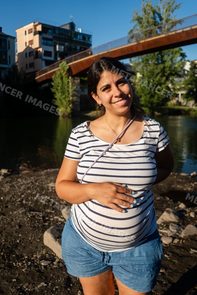 Pregnant woman smiling outdoors by a riverbank with a bridge