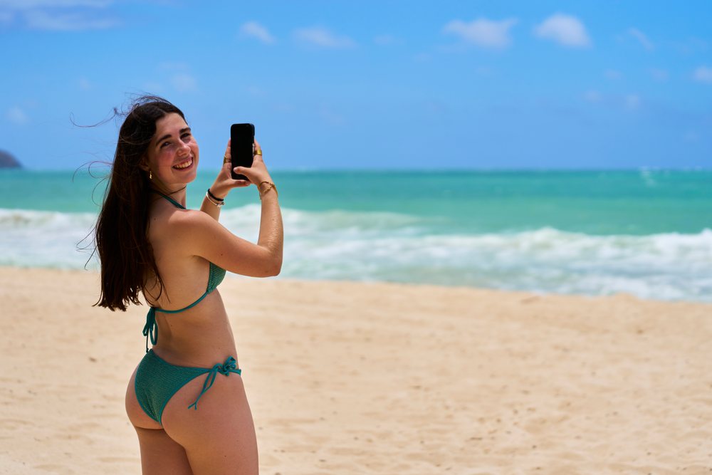 Woman wearing a bikini smiles while taking a photo with a smartphone on a sandy beach by the ocean under a blue sky, Hawaii, USA.