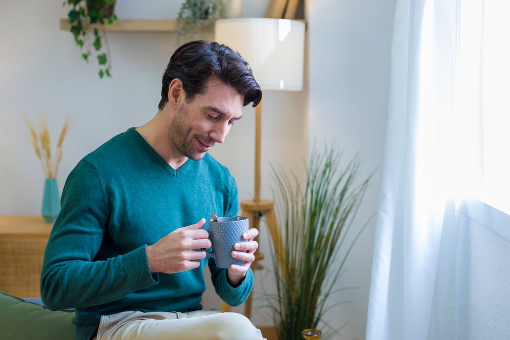 Man in green sweater enjoys coffee seated in a modern home interior with plants and natural light, technology, young adult.