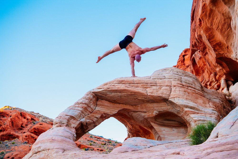 Male performing an acrobatic yoga one hand stand on a natural rock arch in a desert landscape with clear blue sky, USA.