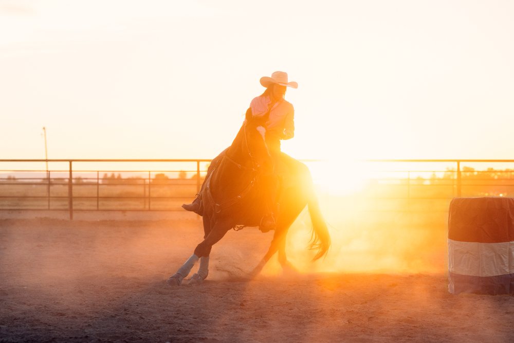 Cowgirl wearing a western hat rides a horse sharply turning around a barrel in an outdoor arena at sunset, Southwest USA.