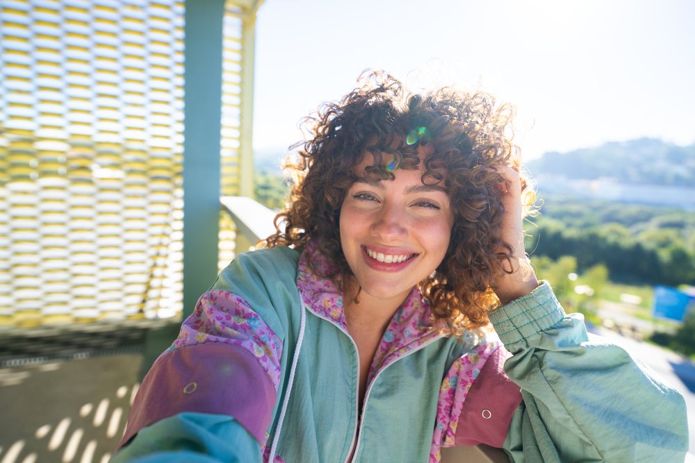 Woman with curly hair smiles while taking a selfie on a sunny outdoor balcony with greenery visible behind her, Spain.