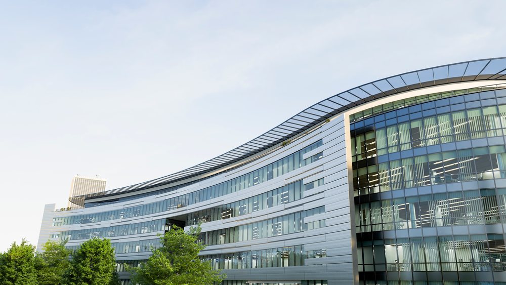 Modern curved office building with metallic facade and large glass windows reflecting sky and trees in daytime urban setting.