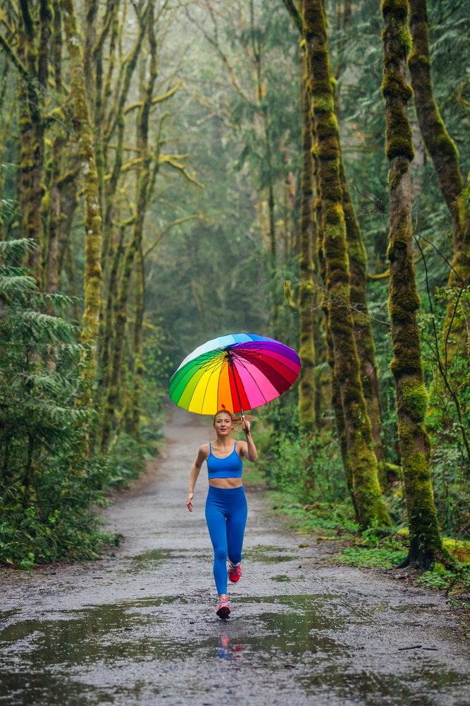 Woman running on a wet forest trail in the USA holding a colorful rainbow umbrella during rainy weather.