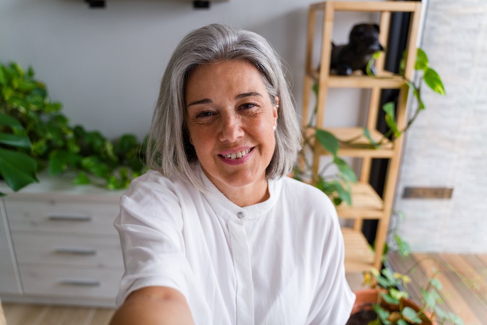 Middle aged woman with gray hair smiling while taking a selfie indoors in a modern home with houseplants, Spain.