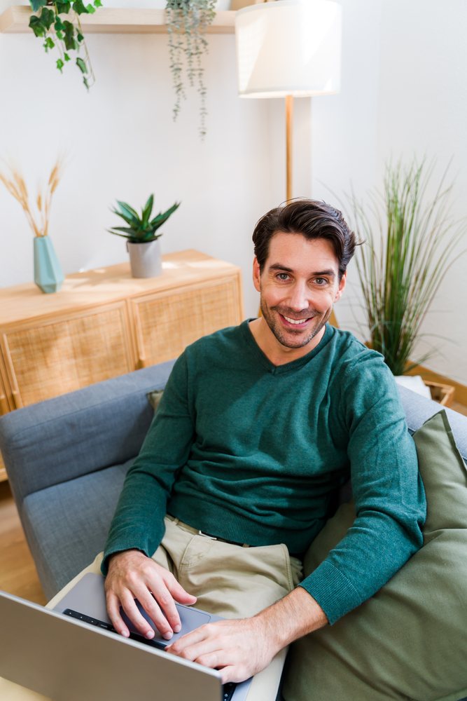 Man sits on a gray sofa using a laptop in a modern living room with plants and wooden furniture, Spain.