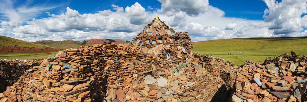 Mani Wall of stacked prayer stones outdoors on grassland with mountains and blue sky at Songge Mani Shijingcheng, China, Dangkuai, Sichuan Province, People's Republic of China.