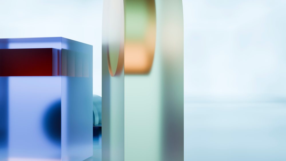 Translucent blue and green geometric glass blocks with smooth reflective surfaces are arranged against a soft, blurred neutral background in a studio setting.