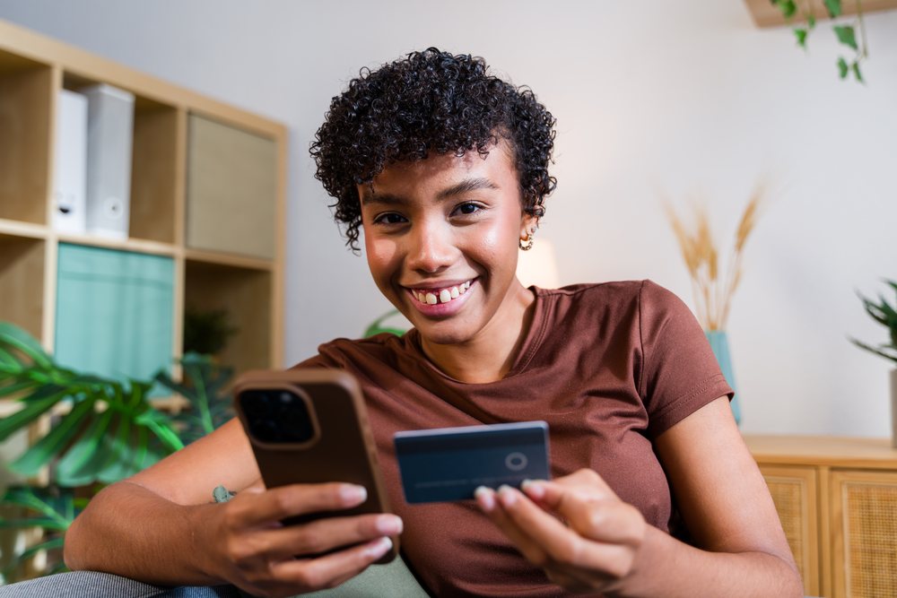 Smiling woman holding a credit card and using a smartphone indoors in a modern home setting, Spain.