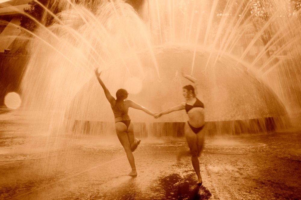 Two women dancing and practicing acrobatics under water spray from a fountain in an outdoor setting, partner, yoga, USA.