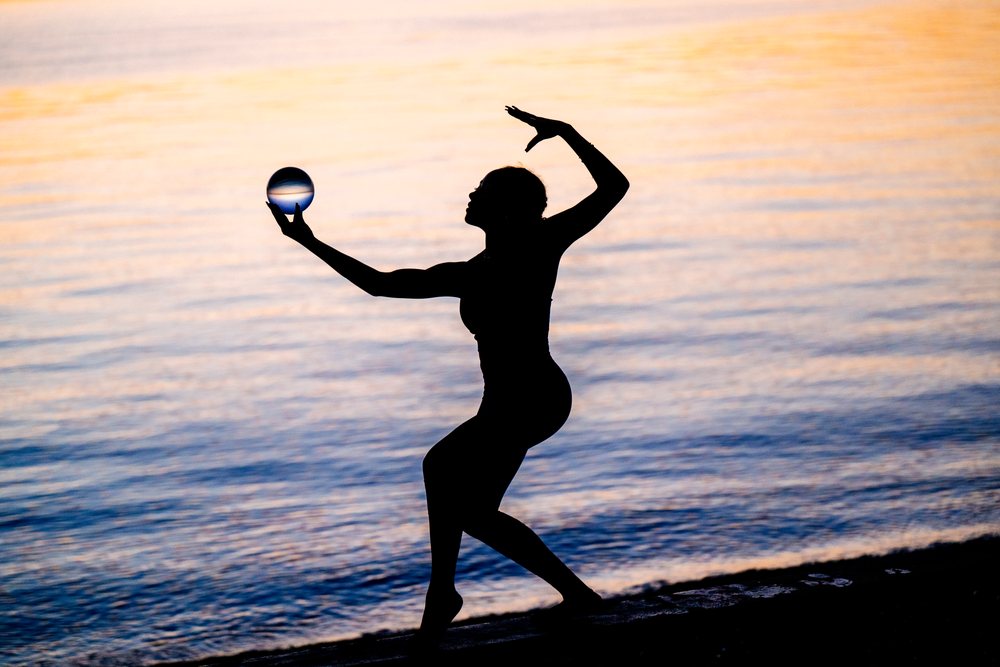 Woman practicing yoga acrobatics balancing a ball on the shore during sunset over water, USA.