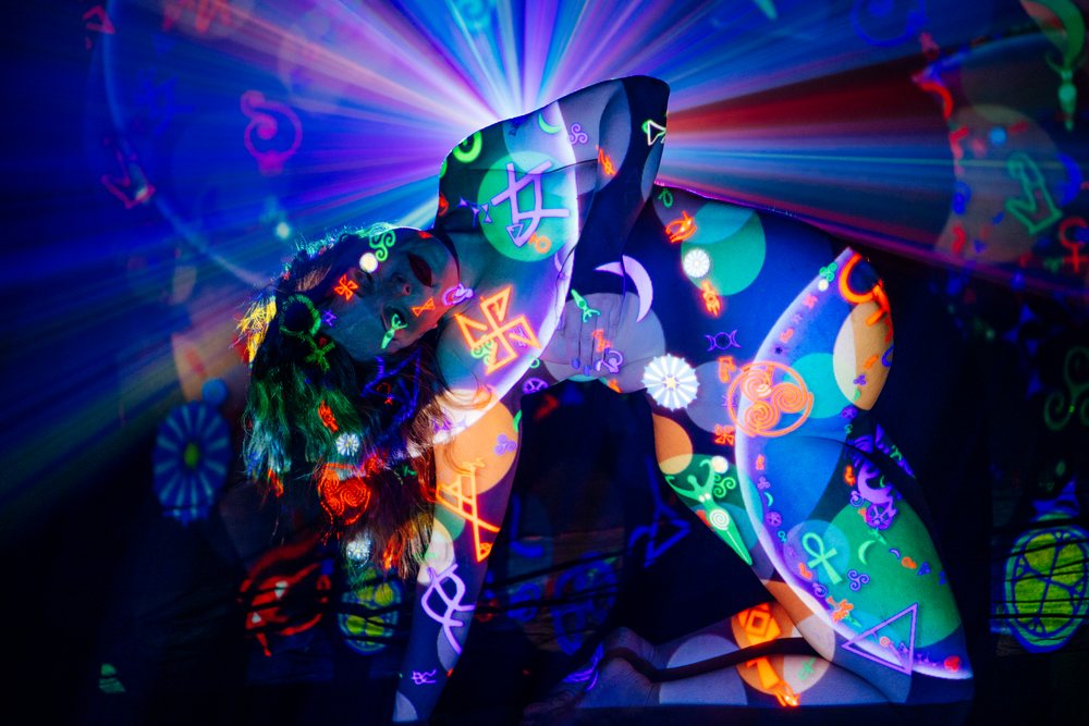 Woman kneels in a dark setting with her body covered by multicolor neon symbol projections and light rays, pregnancy.