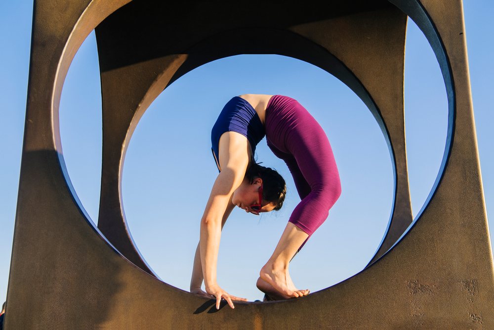 Woman barefoot in purple leggings and blue top performs a backbend yoga pose framed by a circular outdoor metal sculpture under a clear blue sky, USA.