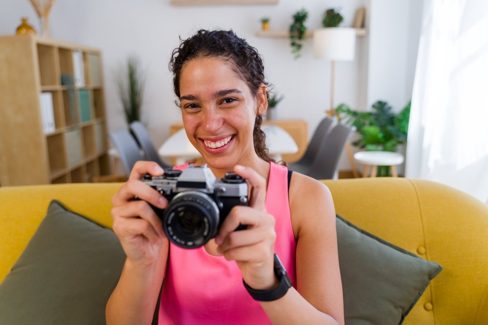 A young woman smiles while holding a vintage camera in a bright living room, Spain.