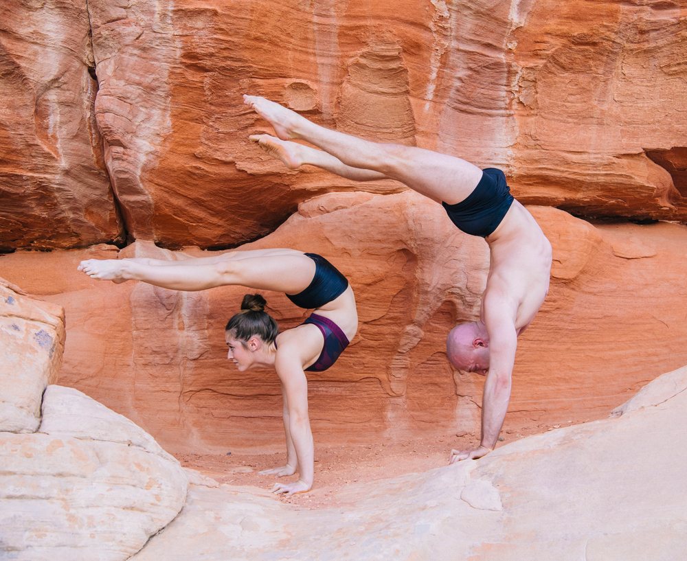 Two people performing yoga handstands balancing on red rock formations outdoors, USA.