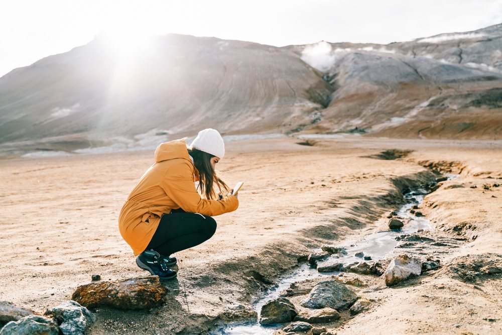 A woman wearing a yellow jacket and white knit hat squats while using her smartphone by a small natural stream in a geothermal area, Iceland.