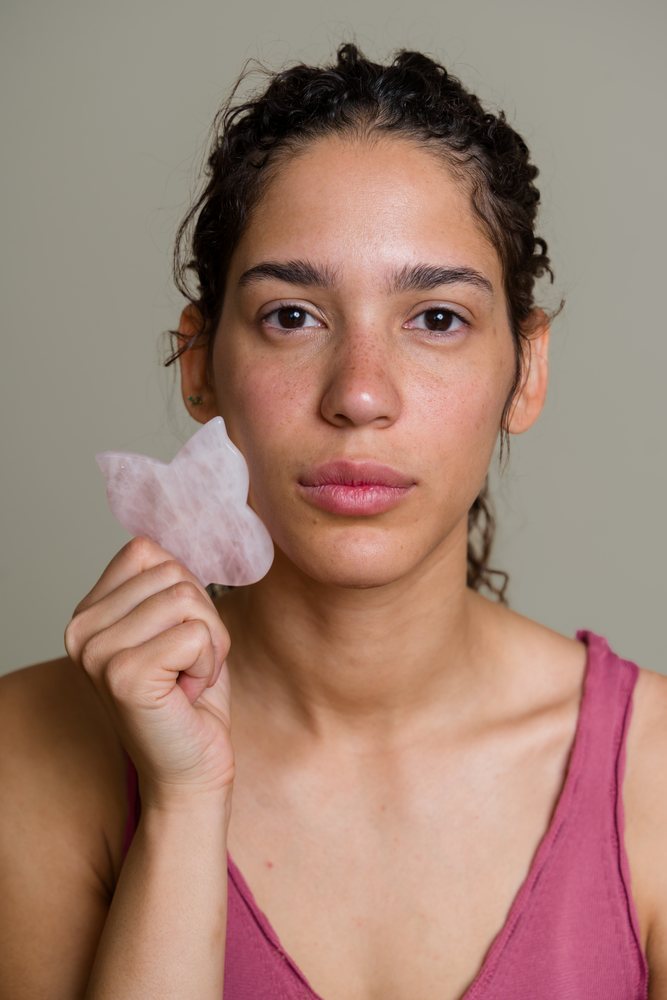 Woman using a rose quartz gua sha tool on her face during a beauty shoot, Spain.