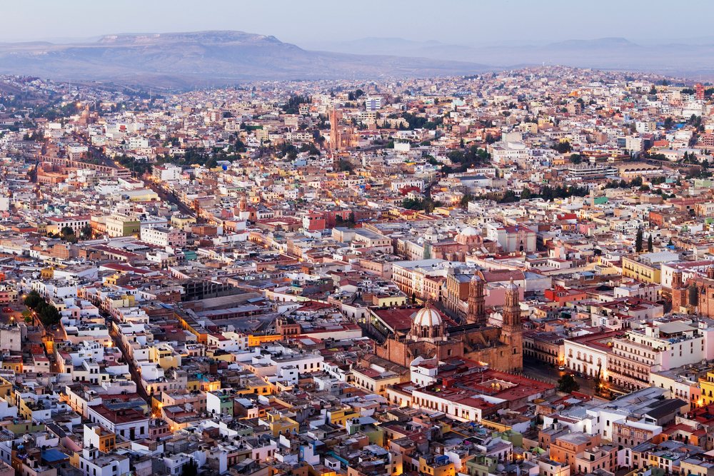 Overlook of Zacatecas city with buildings, streets, dome, twin towers, and mountains under dusk lighting, Mexico.