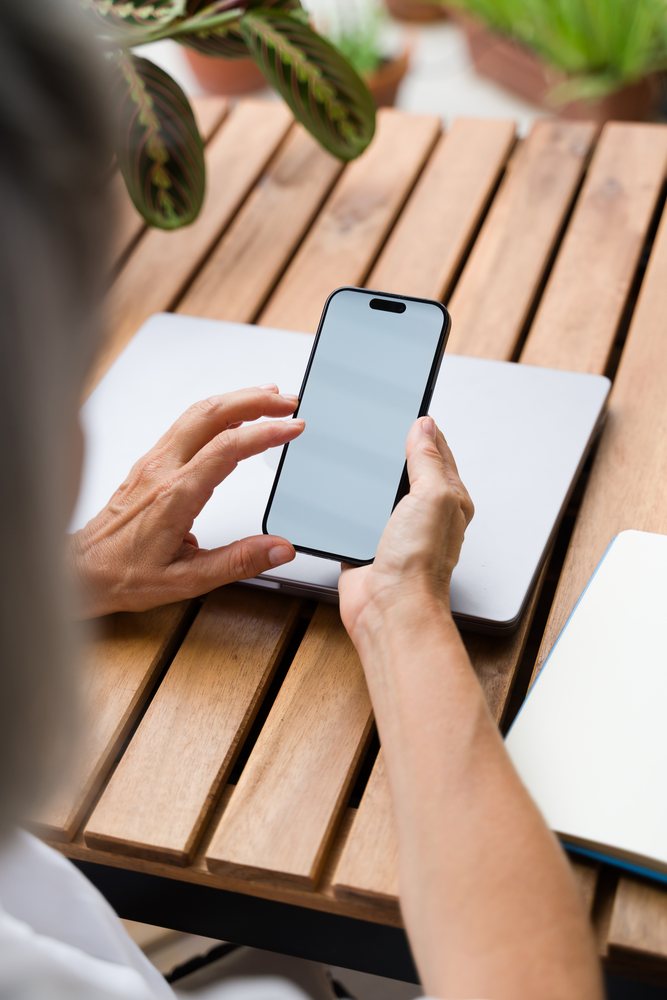 Person holding a smartphone at a wooden table with a closed laptop and notebook, with indoor plants nearby, Spain.