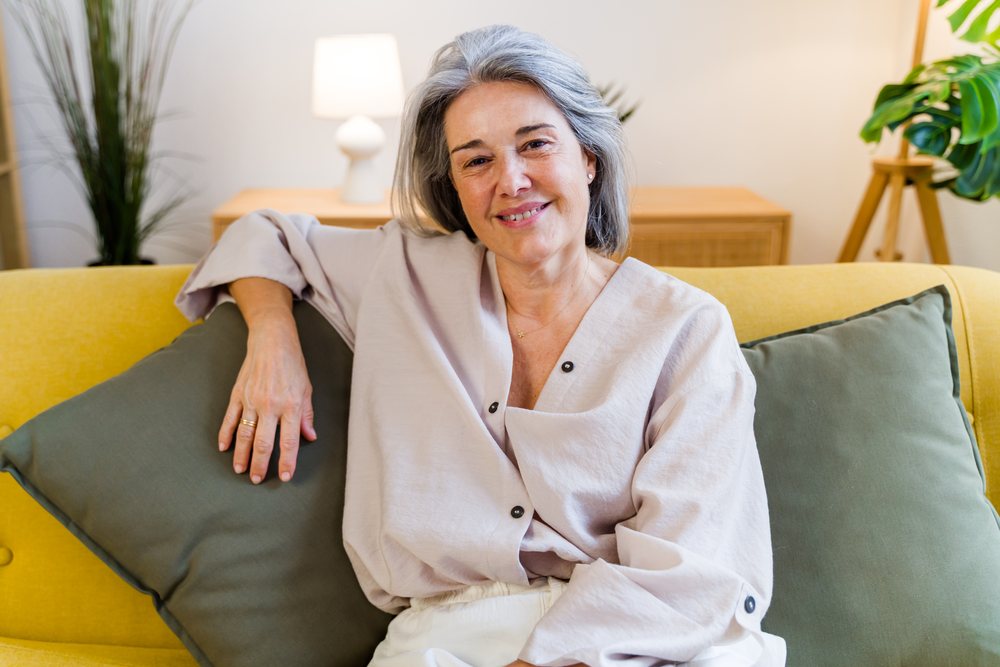 Smiling mature woman with gray hair sits relaxed on a yellow sofa in a cozy home interior, Spain.