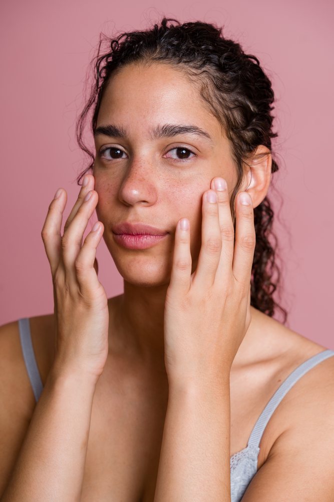 Woman with natural curly hair and freckles touches her face against a pink background, wearing a turquoise sleeveless top, beauty shoot, Spain.