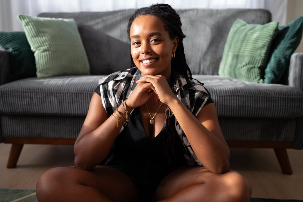 A young woman sits cross-legged on the floor in a living room with a gray sofa and green pillows, moving, Spain.