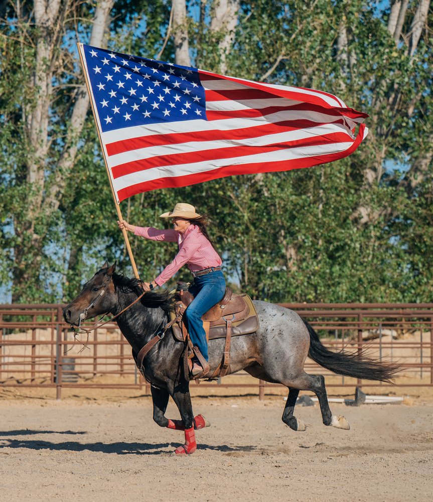 Woman wearing a cowboy hat rides a horse with a saddle and leg wraps while waving an American flag in a dusty outdoor arena, Southwest USA.