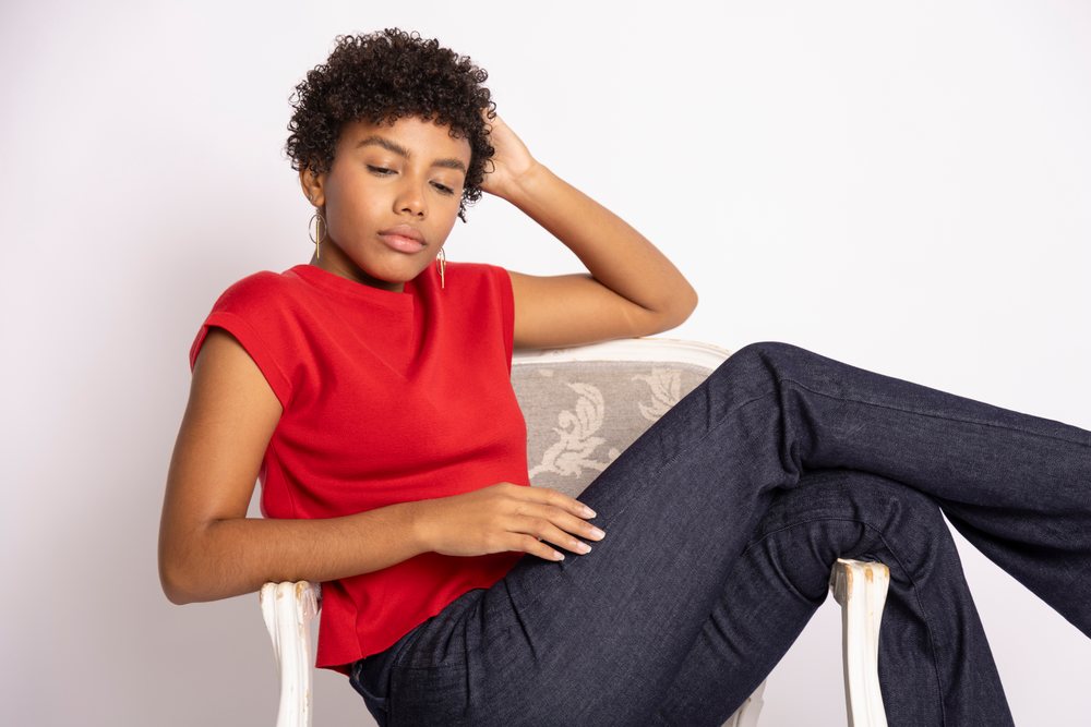 Woman with curly hair wearing a red top and dark jeans sitting relaxed in an armchair in a studio, fashion, clothes, young culture.