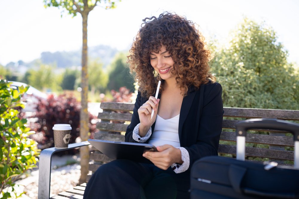 Woman working on digital tablet sitting on park bench outdoors, Spain.