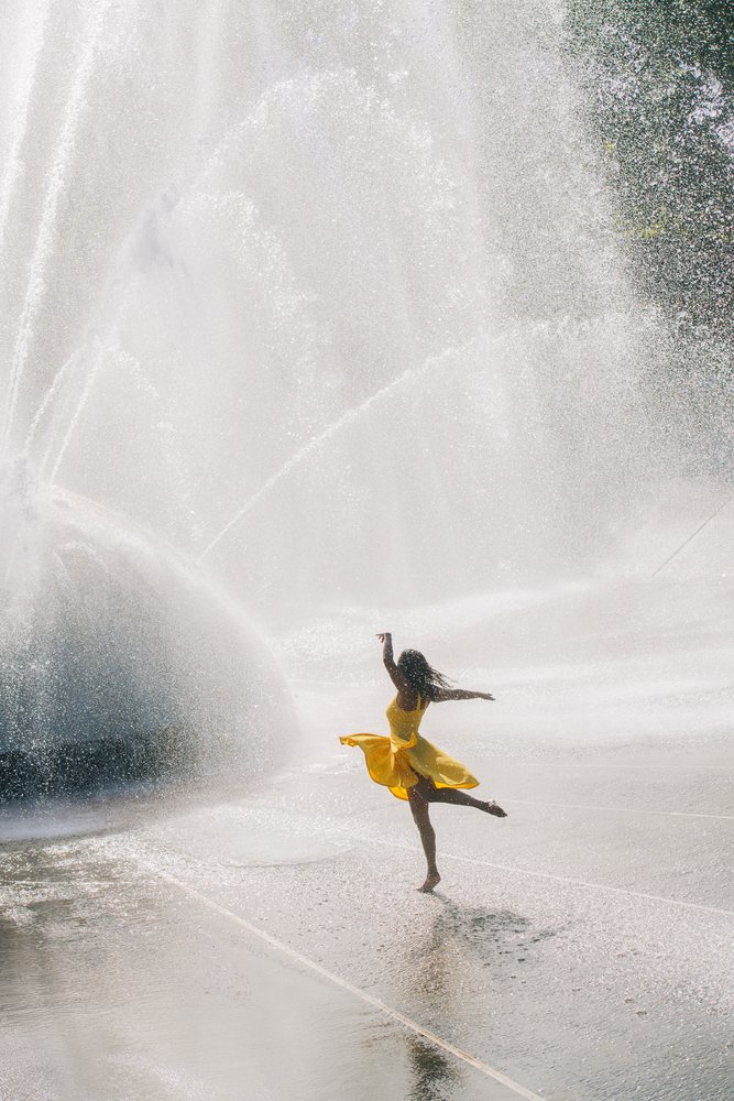Woman in a yellow dress dances joyfully through the water spray of a large outdoor fountain in the USA on a sunny summer day.