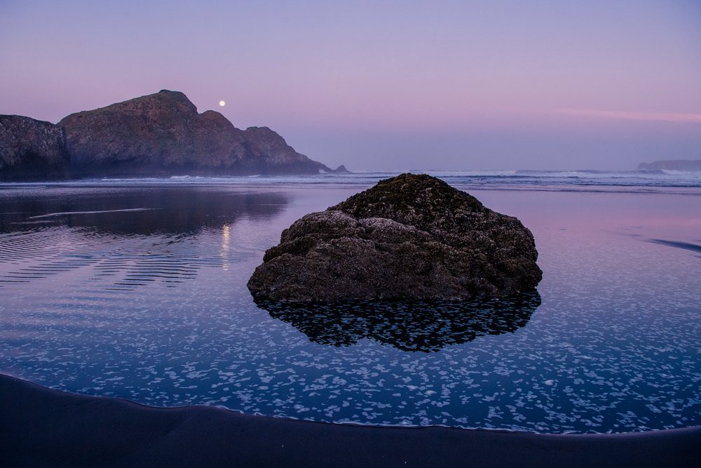 Large rock formation partially submerged in calm coastal water at sunset reflecting moonlight near a mountain at Meyers Creek, Oregon, USA.