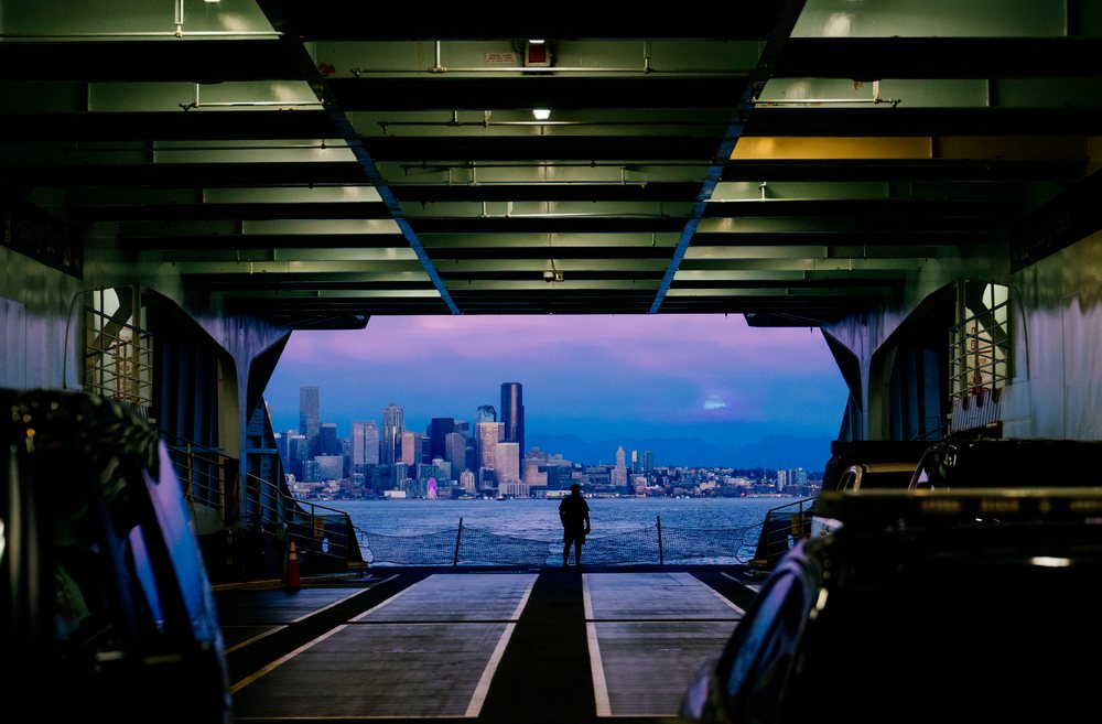 Silhouette of a person standing on a ferry deck with cars, looking at a city skyline across the water at dusk, USA.