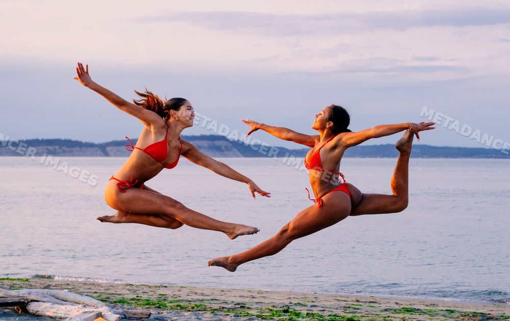 Two women perform acrobatic jumps on a beach at twilight in WA, USA