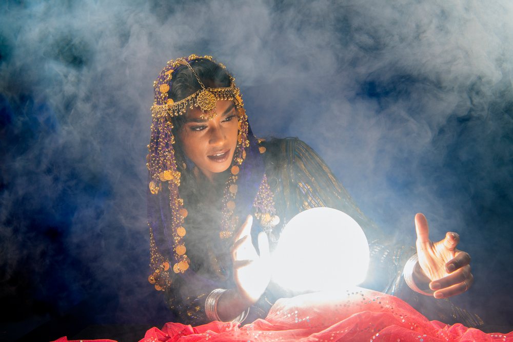 A woman dressed as a fortune teller gazes into a glowing crystal ball surrounded by smoke, wearing gold jewelry and a purple veil in the, USA.