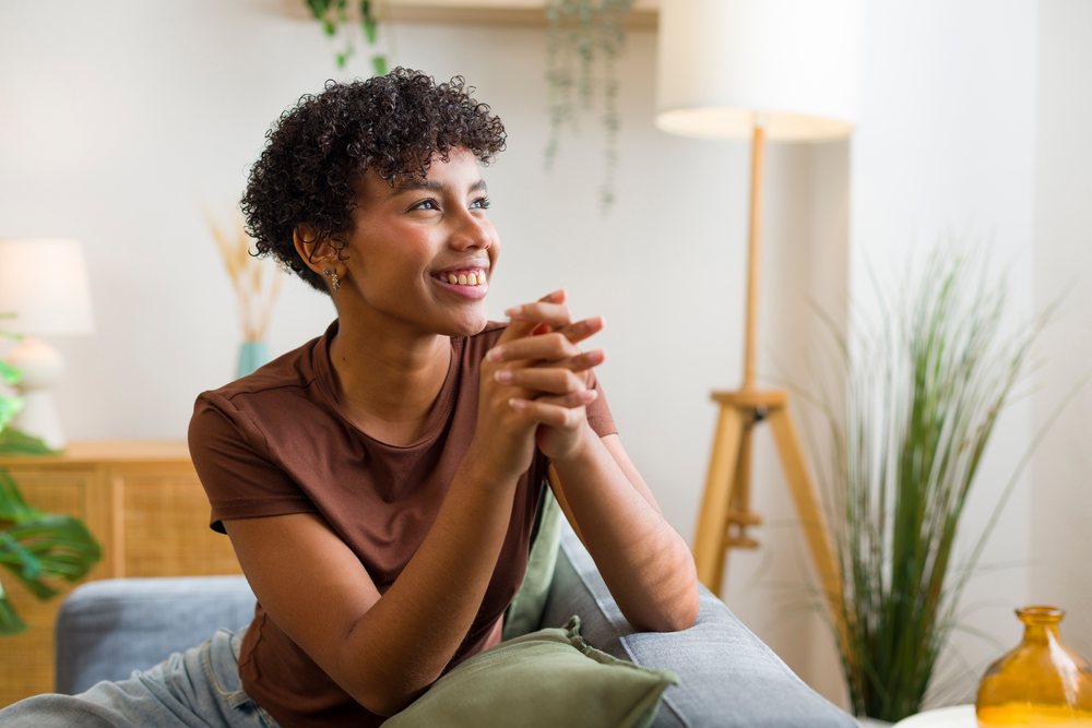 Young woman with curly hair smiling while sitting on a gray sofa in a cozy modern living room interior.