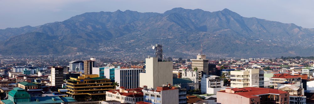 Cityscape of San Jose, Costa Rica showing urban buildings with a mountainous landscape in the background under a cloudy sky.